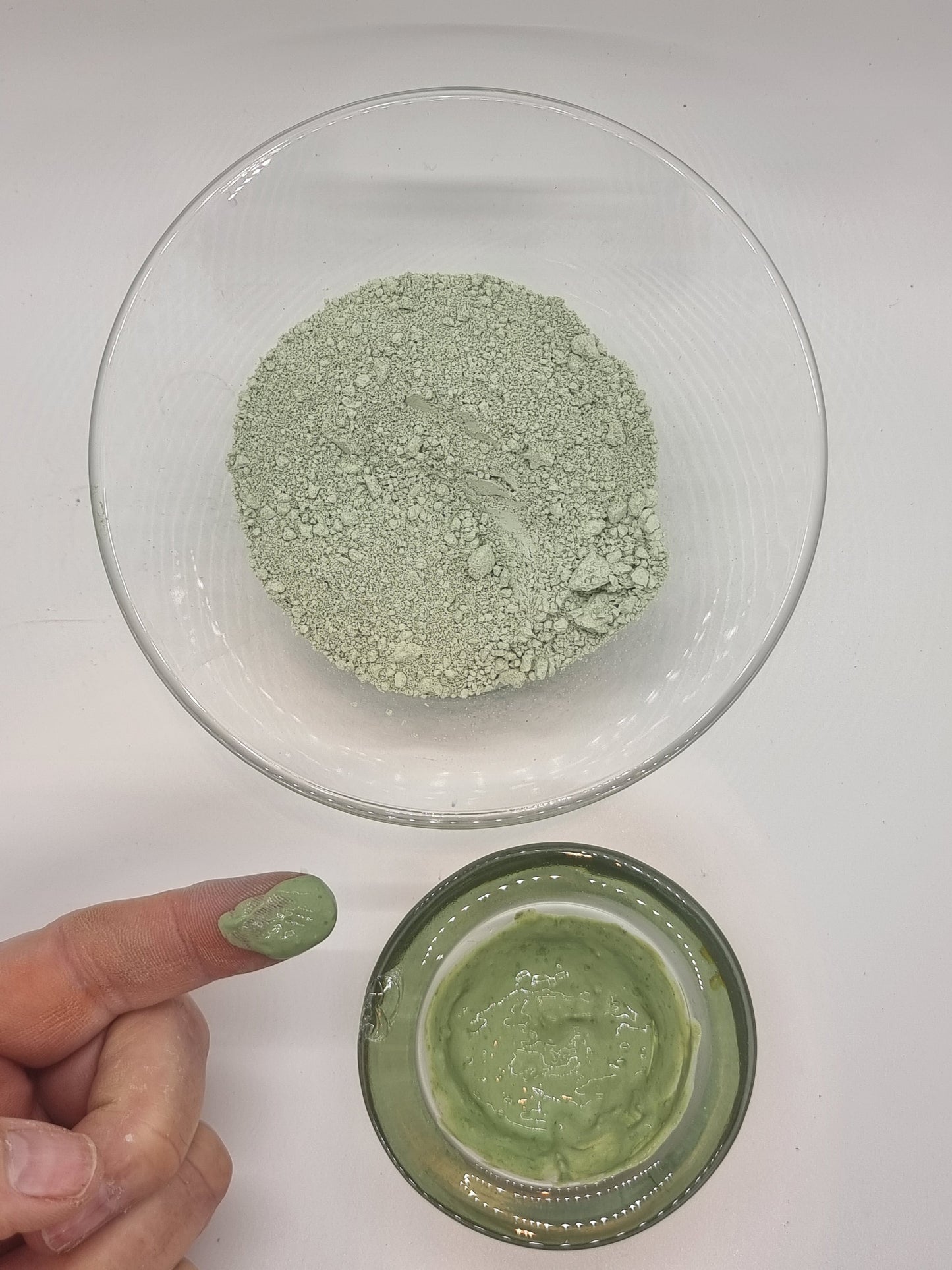 two glass bowls one containing dry fine french green clay that is a mid green colour, the other containing wet green clay that has a shiny appearance. A hand and finger can be seen. The finger has a blob of shiny wet clay on it that is also green in colour.