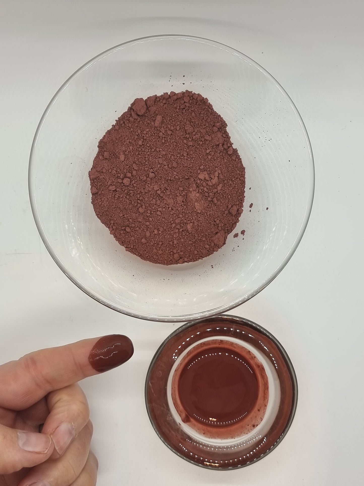 two glass bowls one containing deep earthy red Moroccan Ghassoul/Rhassoul clay. One bowl contains dry clay the other a paste of clay and water which looks shiny. There is a hand and a finger showing that has been dipped into the wet clay some of which sits on the end of the finger with the light reflecting off it.