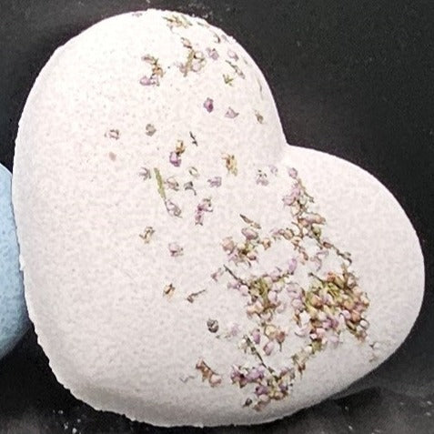 Heart shaped bath bomb in a light purple pink colour made using brazilian purple clay from Bath and Body Base limited. Decorated with small green and purple heather flowers