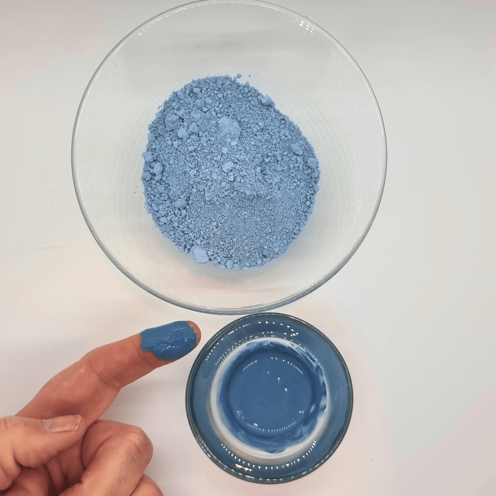 Serbian Blue Clay in glass bowls, one dry one wet, with a hand dipped into the wet clay showing the vibrant natural colour