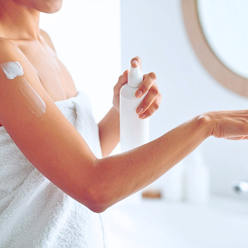 woman in a white towel applying body lotion to her arm from a pump bottle. Bathroom mirror in background