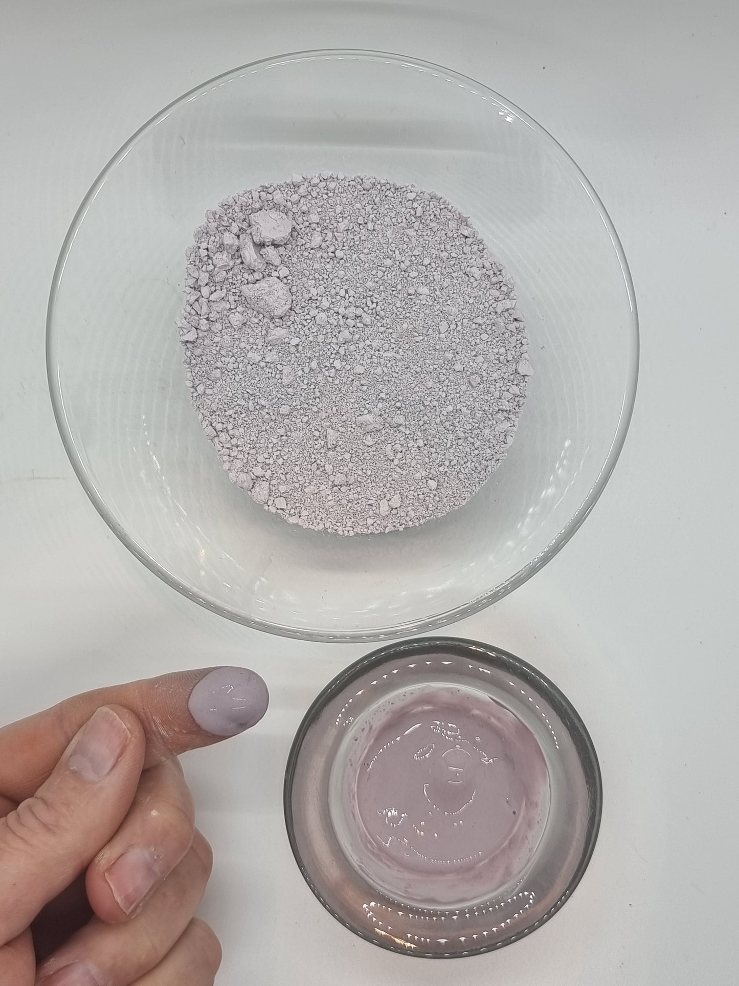 two glass bowls one containing fine brazilian purple clay dry powder, the other purple clay paste. A hand is visible with a finger that has a blob of wet purple clay on the end.